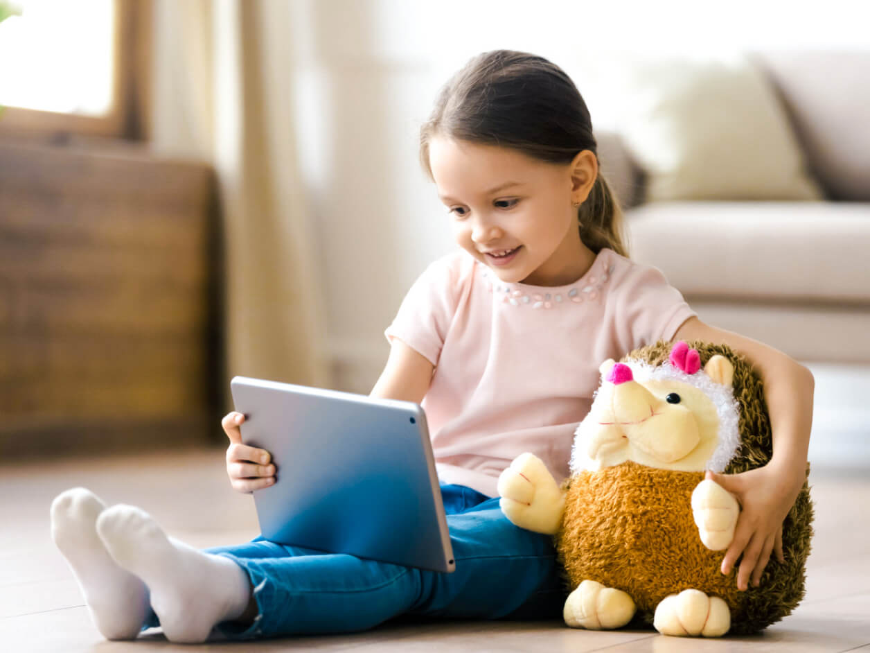 A small child happily plays with a tablet device while holding a stuffed animal.