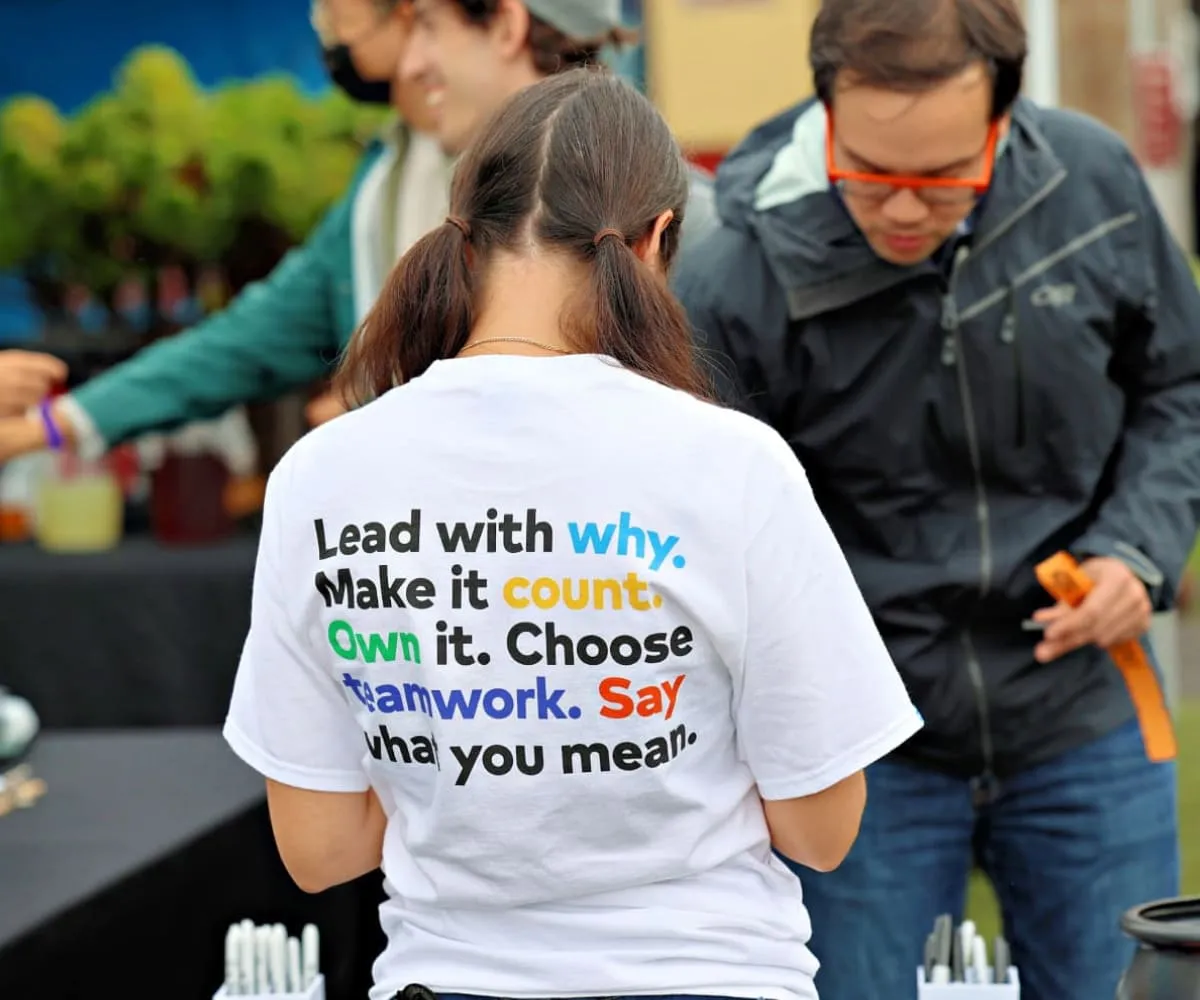 A female Thumbtack employee with her back to the camera. The back of her shirt reads out the Thumbtack values.