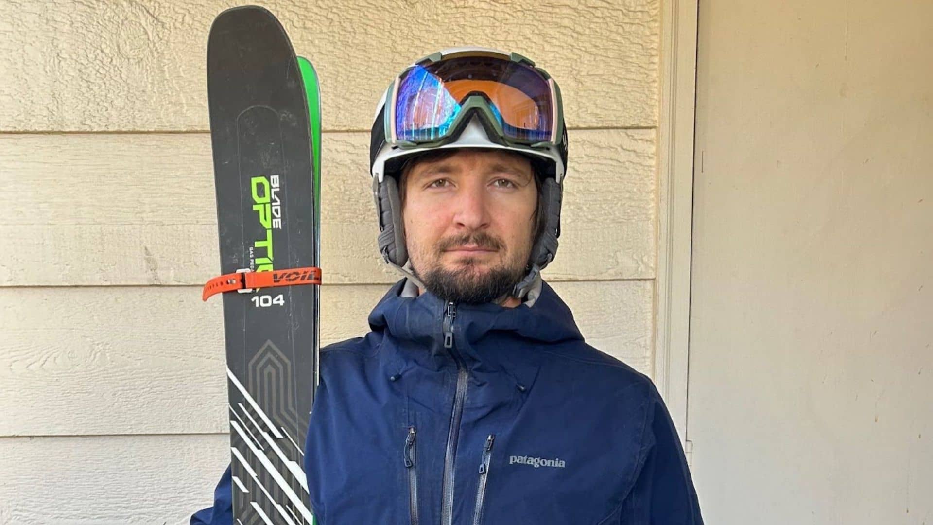 A skier stands with a long pair of skis next to him in front of a beige wall