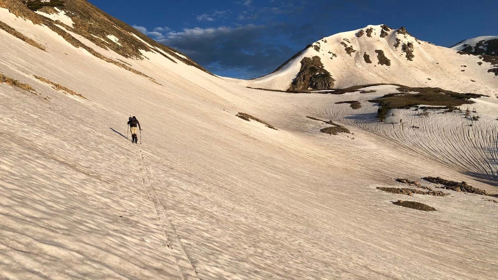 A skier heads up a hill with the aid of backcountry ski bindings. There's a large snowy mountain in the background, bathed in morning light