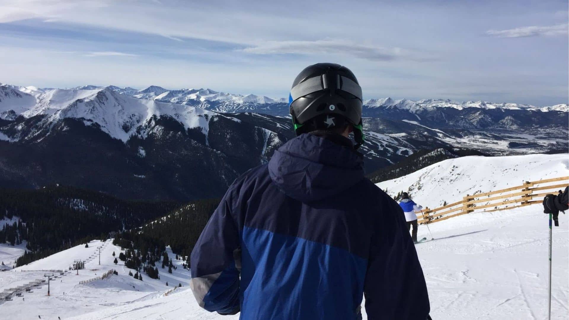 A skier prepares to descend a slope with a helmet on and mountains in the background