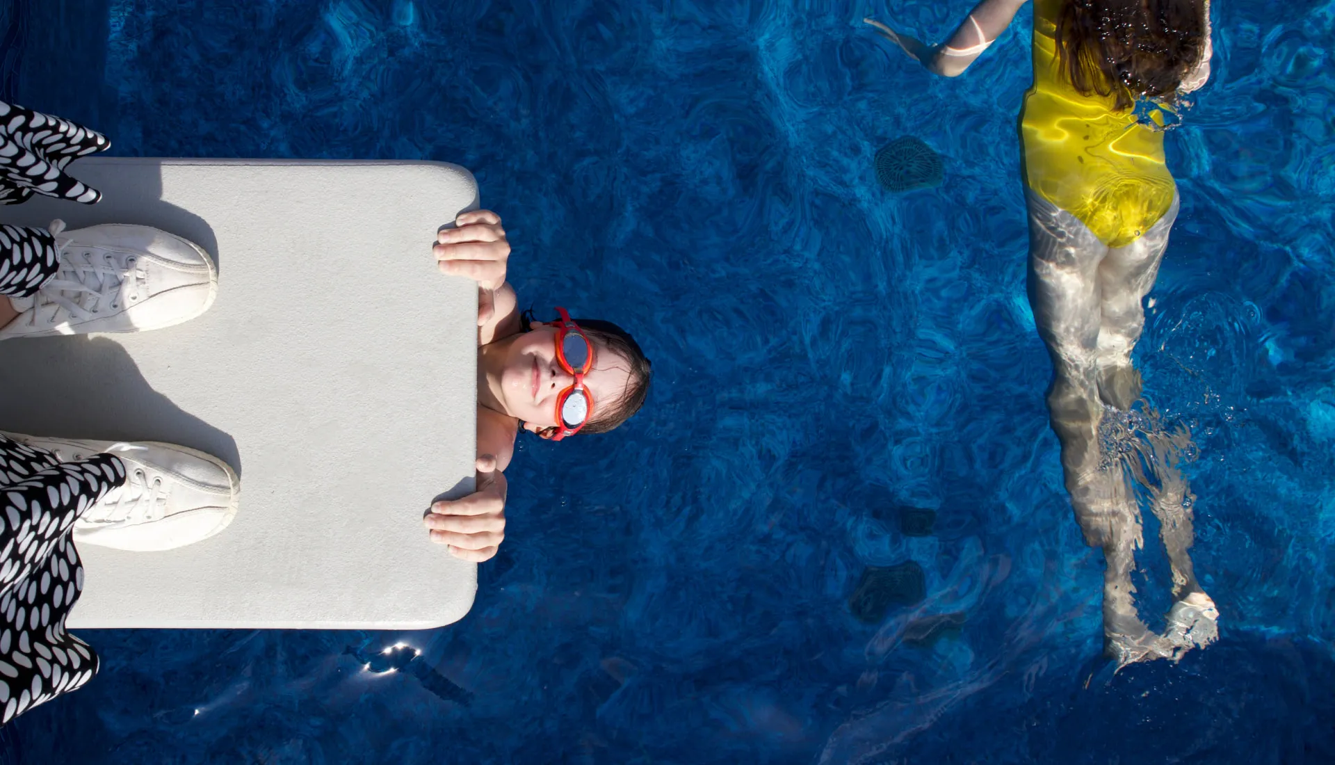 Kid with goggles hanging off diving board in swimming pool as person in yellow bathing suit swims by