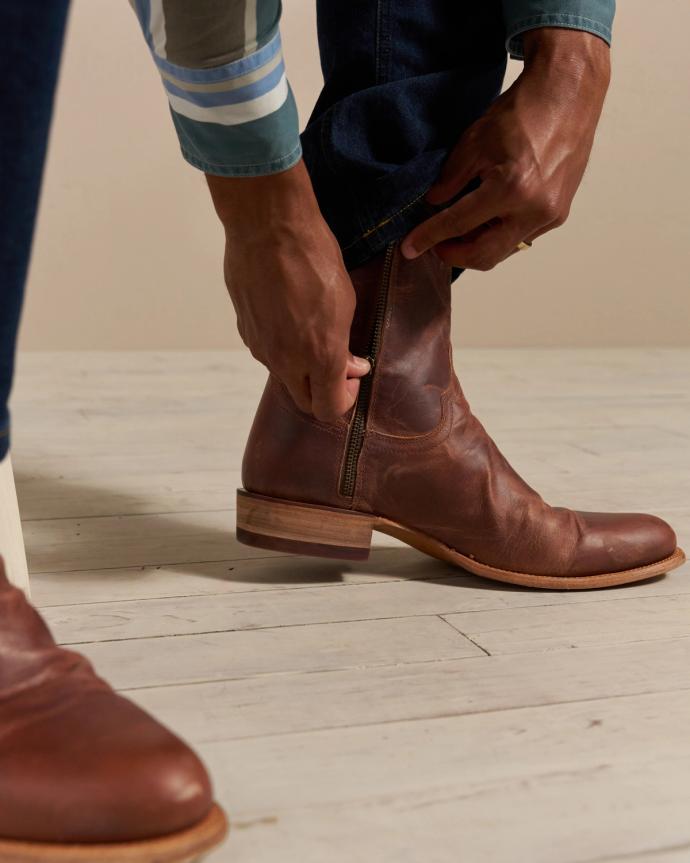 A person wearing jeans and a long-sleeved shirt is zipping up a brown leather boot. Only the person's hands, feet, and part of their legs are visible.