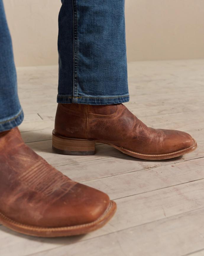 Close-up of a person wearing worn brown leather boots and blue jeans standing on a wooden floor.