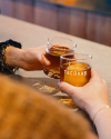 Two hands clinking glasses filled with amber liquid, one glass branded with "tecovas," over a wooden table.