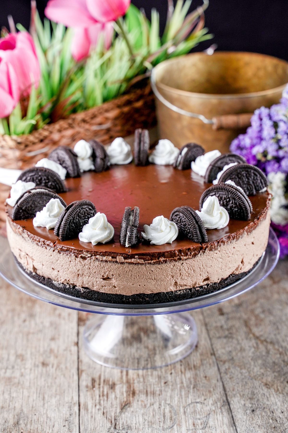 cheesecake on glass cake stand with basket of flowers in the background