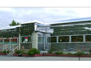 View of front and entrance to diner. It is an old-fashioned stainless steel diner exterior with many windows, like a train car.