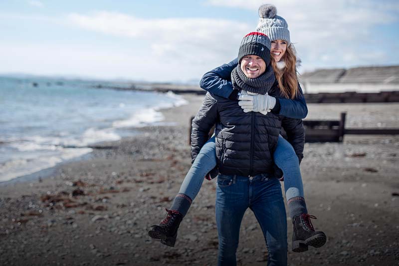 Couple wearing Heat Holders piggyback on a beach