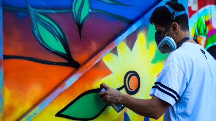 A man wearing a full face mask spray-painting a mural of a flower.