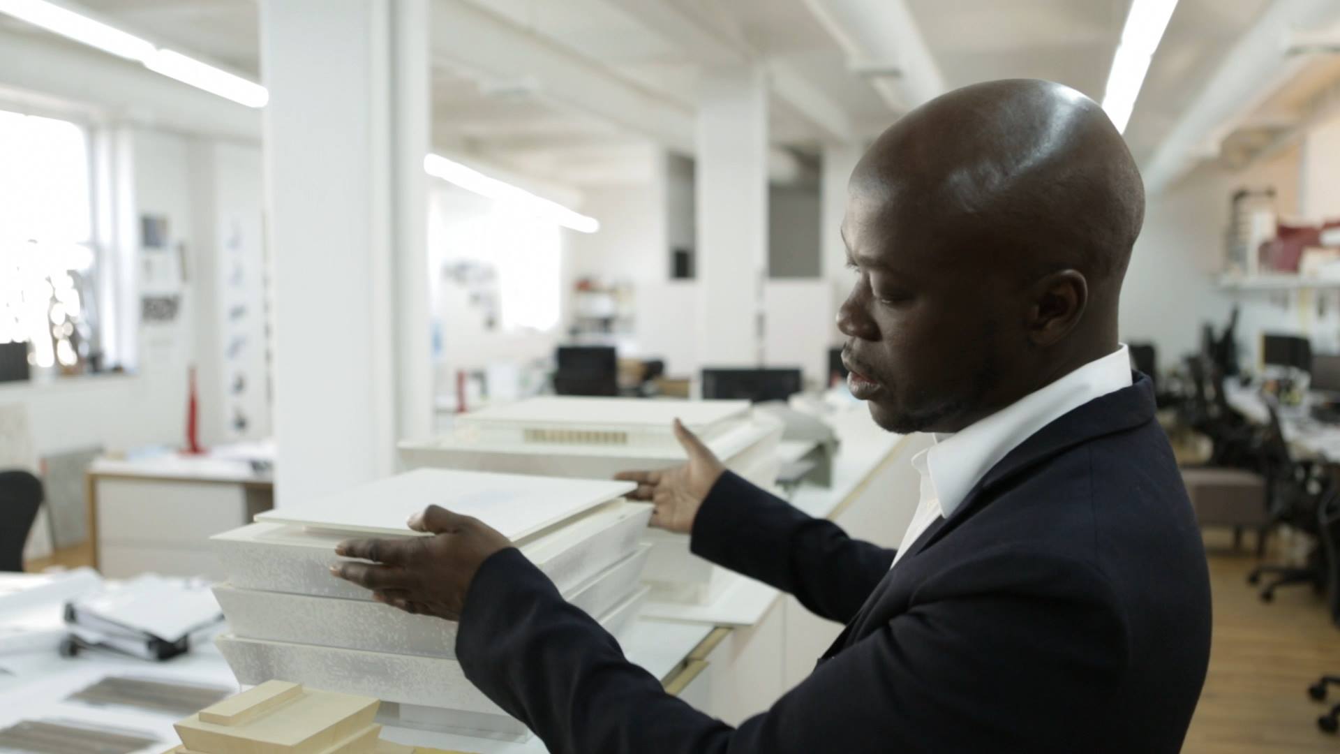 David Adjaye arranging a stack of white blocks on a table with offices in background