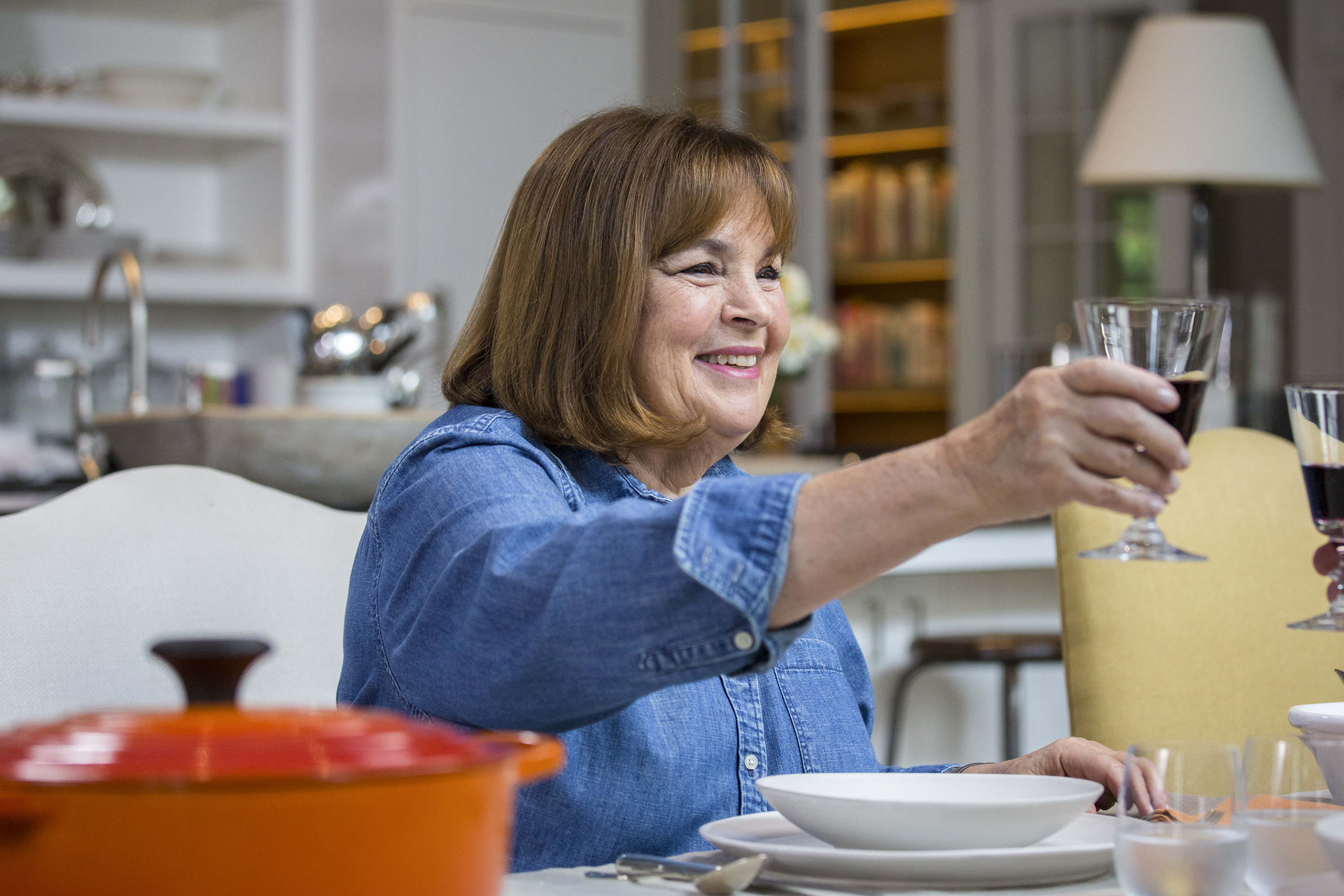 A woman, Ina Garten, wearing a denim shirt and seated at a table raises a glass.