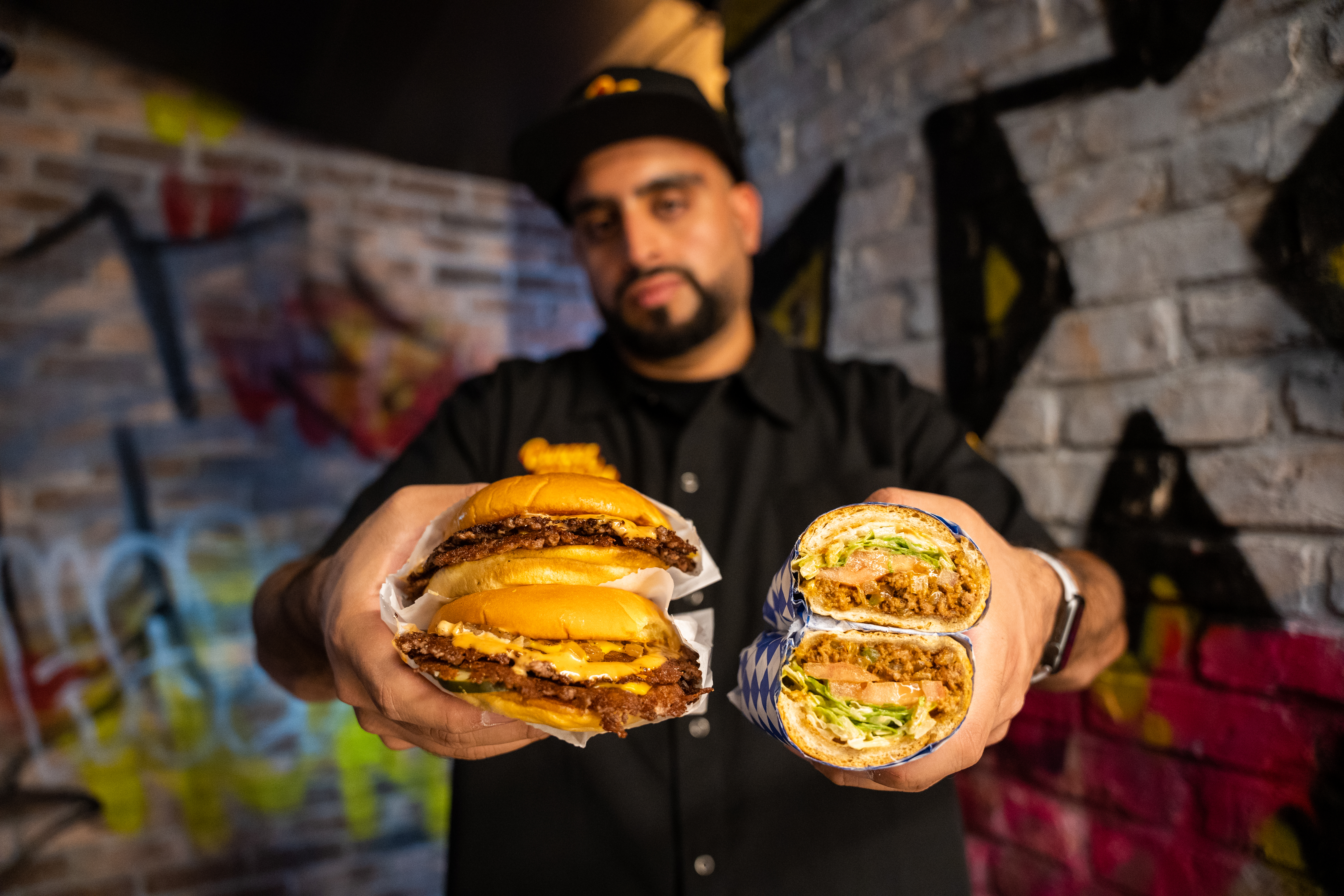 Burger Bodega Abbas Dhanani holding a smash burger and chopped cheese sandwich in front of a graffiti-ed wall.