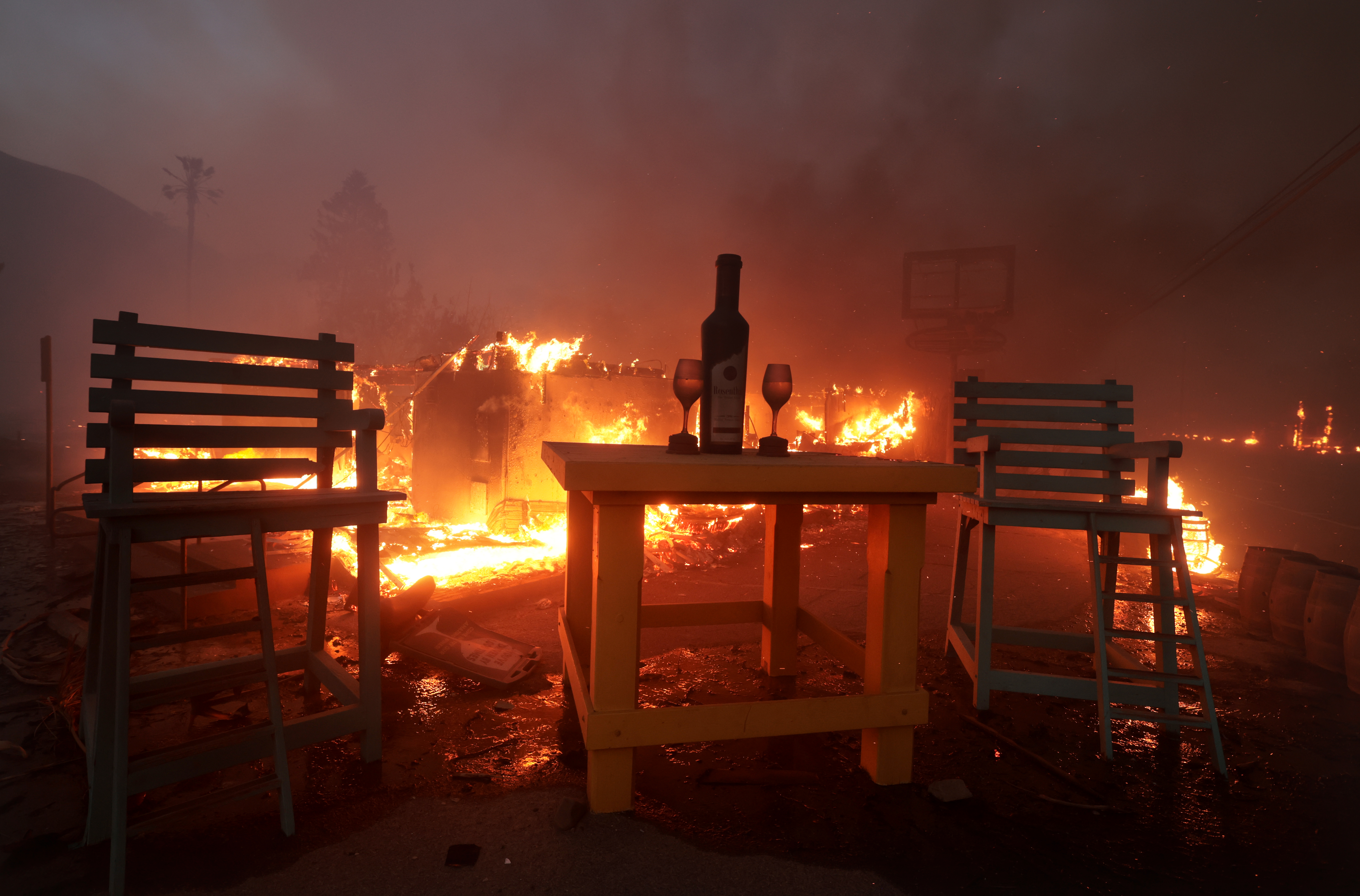 A fire persists outside of a Malibu wine bar called Rosenthal with a wine bottle and glasses on a table remaining.