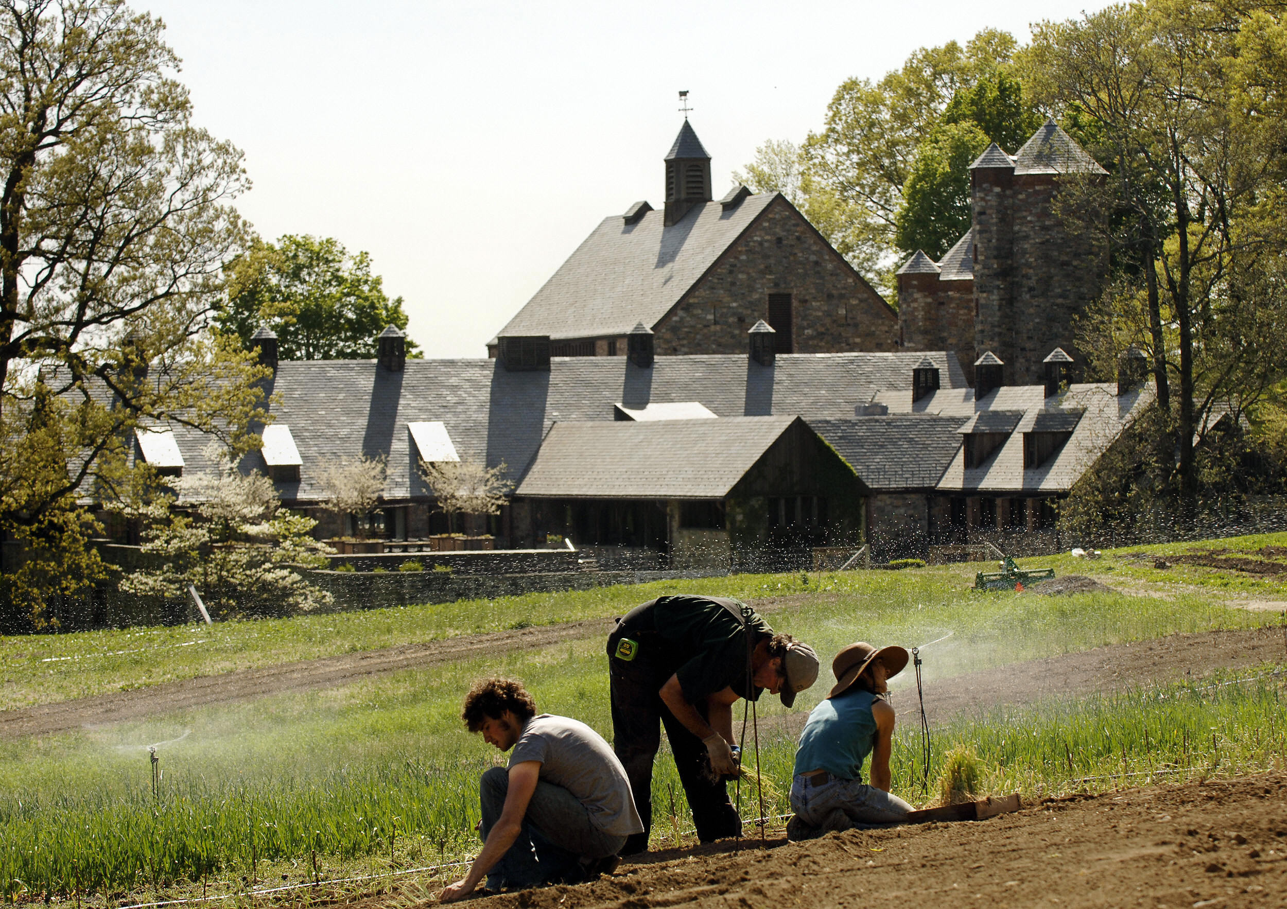 In the foreground, three people appear to garden as sprinklers run. In the background there is a large stone building with pitched roofs surrounded by trees.