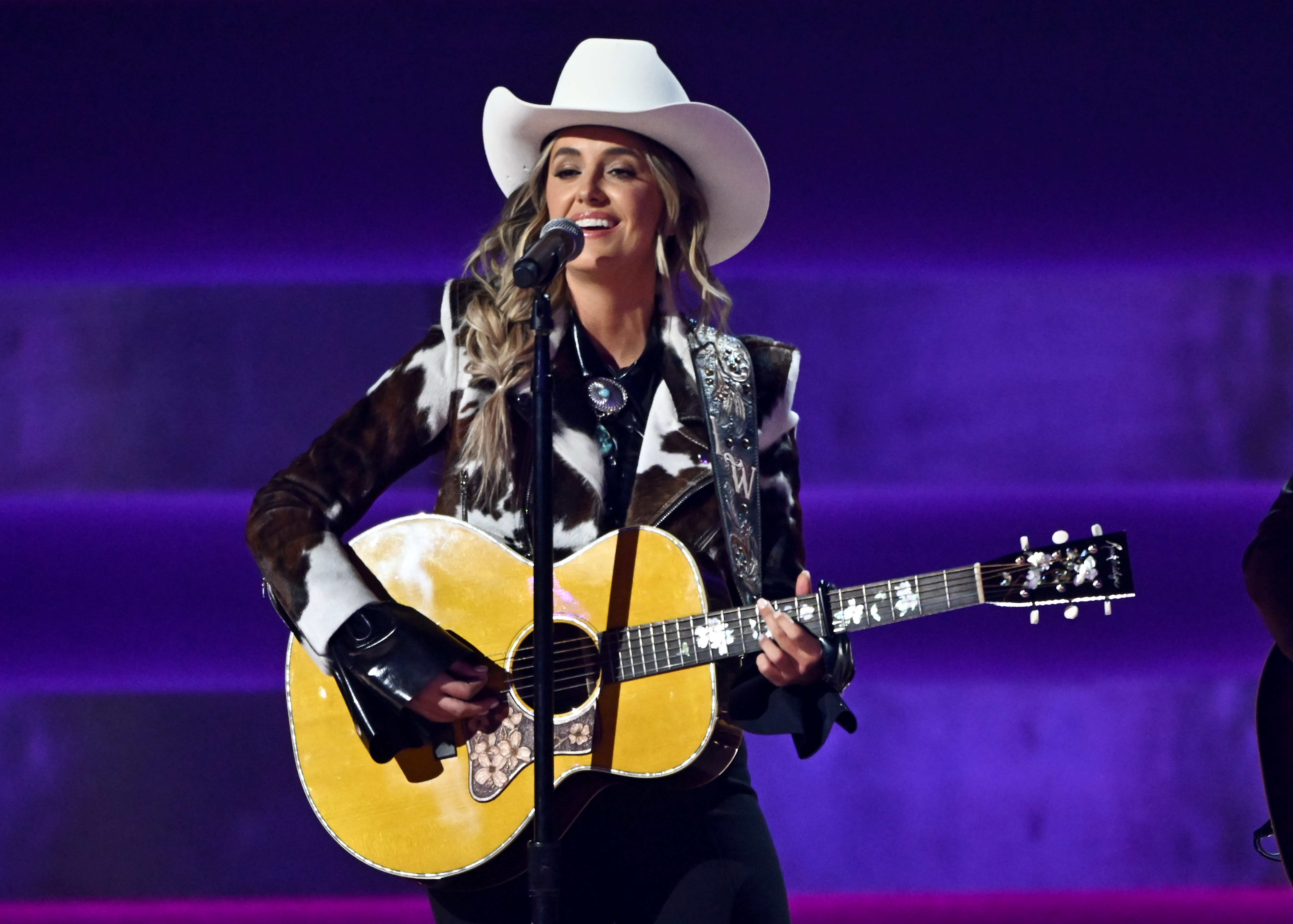 Lainey Wilson performs in a Nudie-esque suit and white cowboy hat, while strumming a yellow acoustic guitars at the 58th Annual CMA Awards.