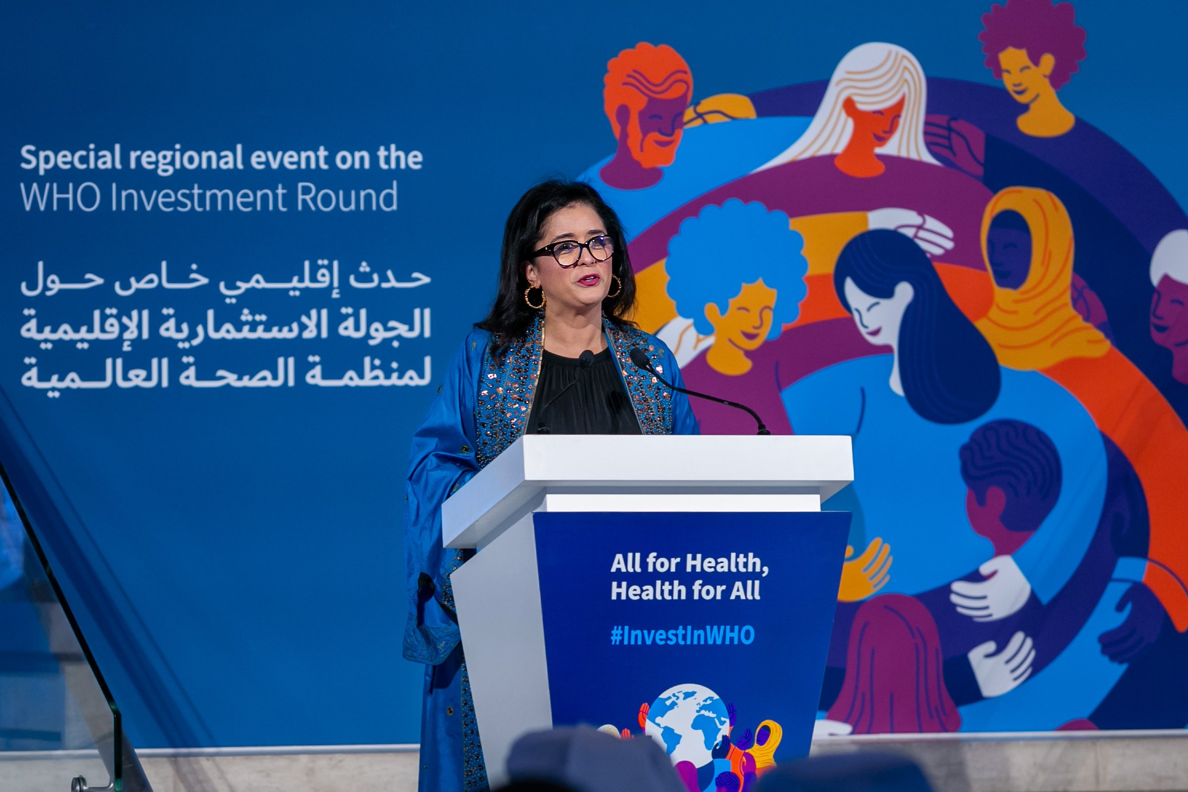 A woman at a speaker podium in front of a dark blue backdrop with a colorful illustration of people embracing each other and the text "Special regional event on the WHO Investment Round".