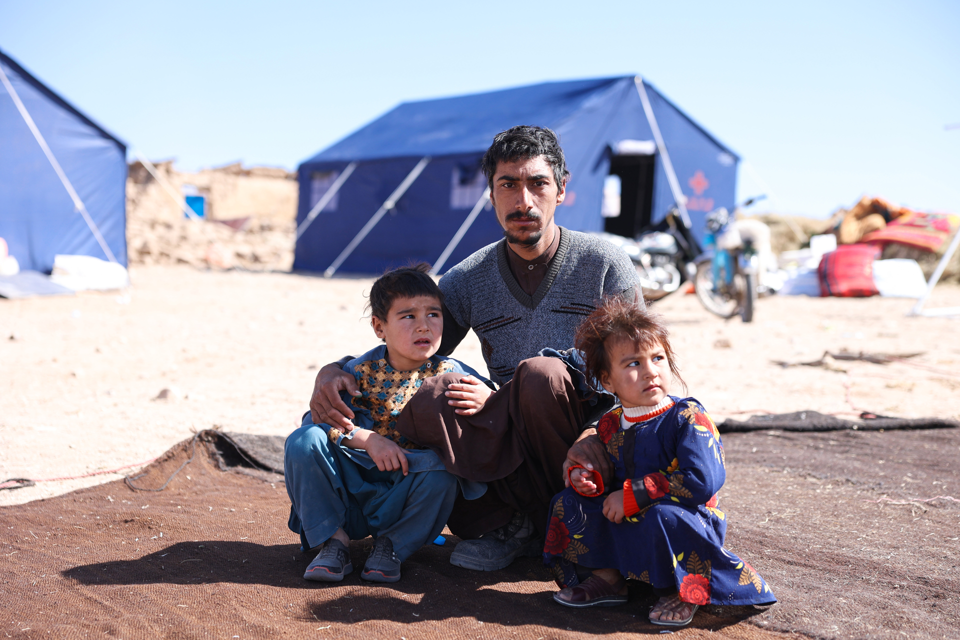 A man and a young girl and boy with worried expressions on their faces sit on a mat in front of large tents