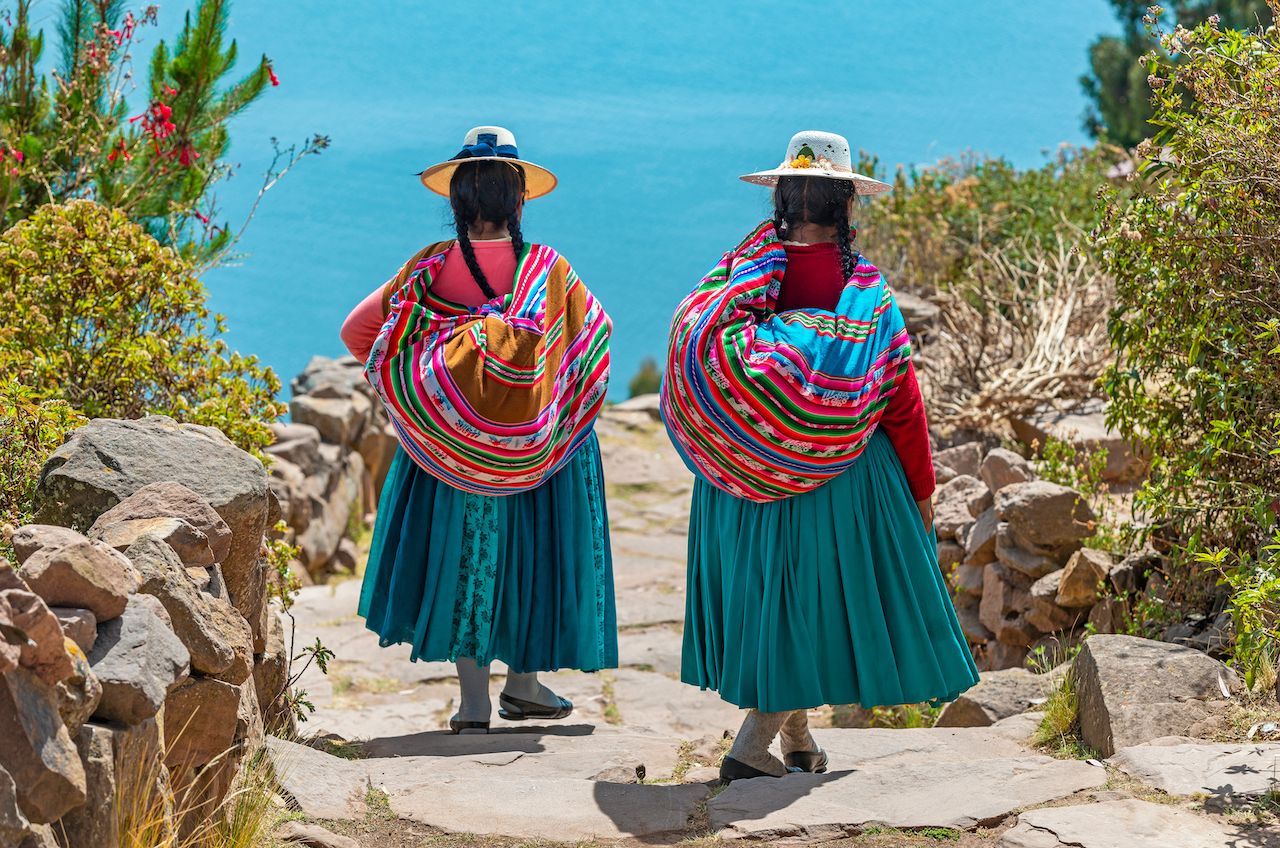 Two indigenous Quechua women in traditional clothes walking down the path to the harbor of Isla Taquile (Taquile Island) with the Titicaca Lake in the background, Peru., Uros Islands