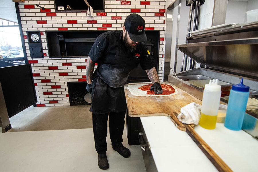 A man in flour-covered black clothing, including Sally's Apizza branded hat and shirt, spreads tomato sauce on pizza dough.