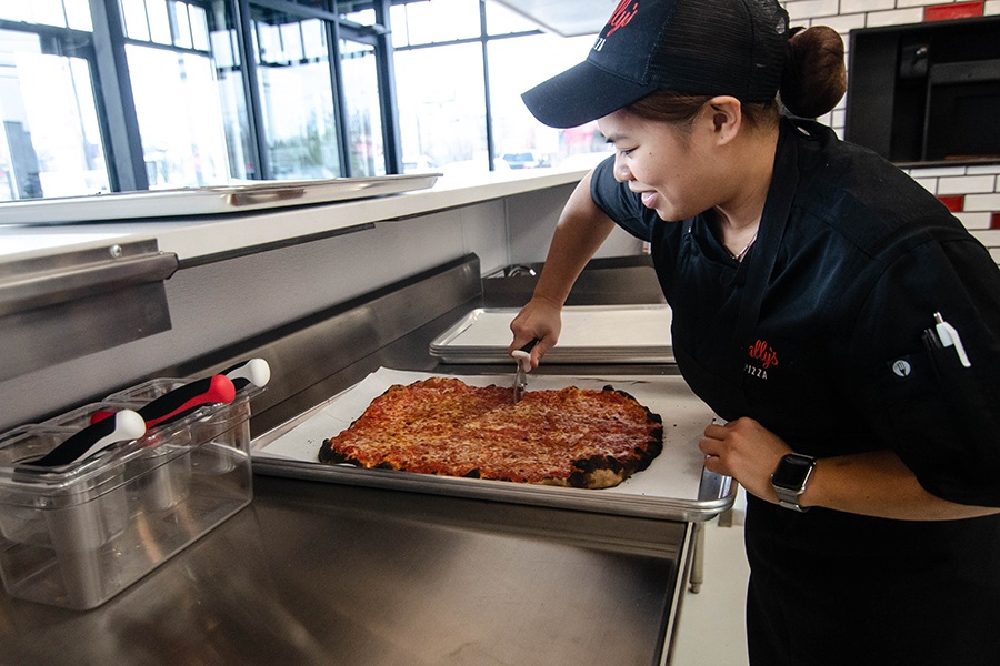 A woman in Sally's Apizza branded hat and shirt slices of a cheese pizza on a metal counter.