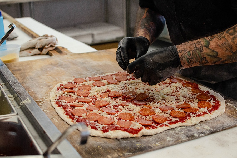 Gloved hands put pepperonis on a thin-crust uncooked pizza.