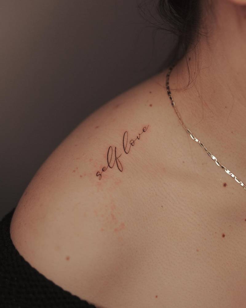 "Self love" lettering tattoo placed on the top of shoulder.