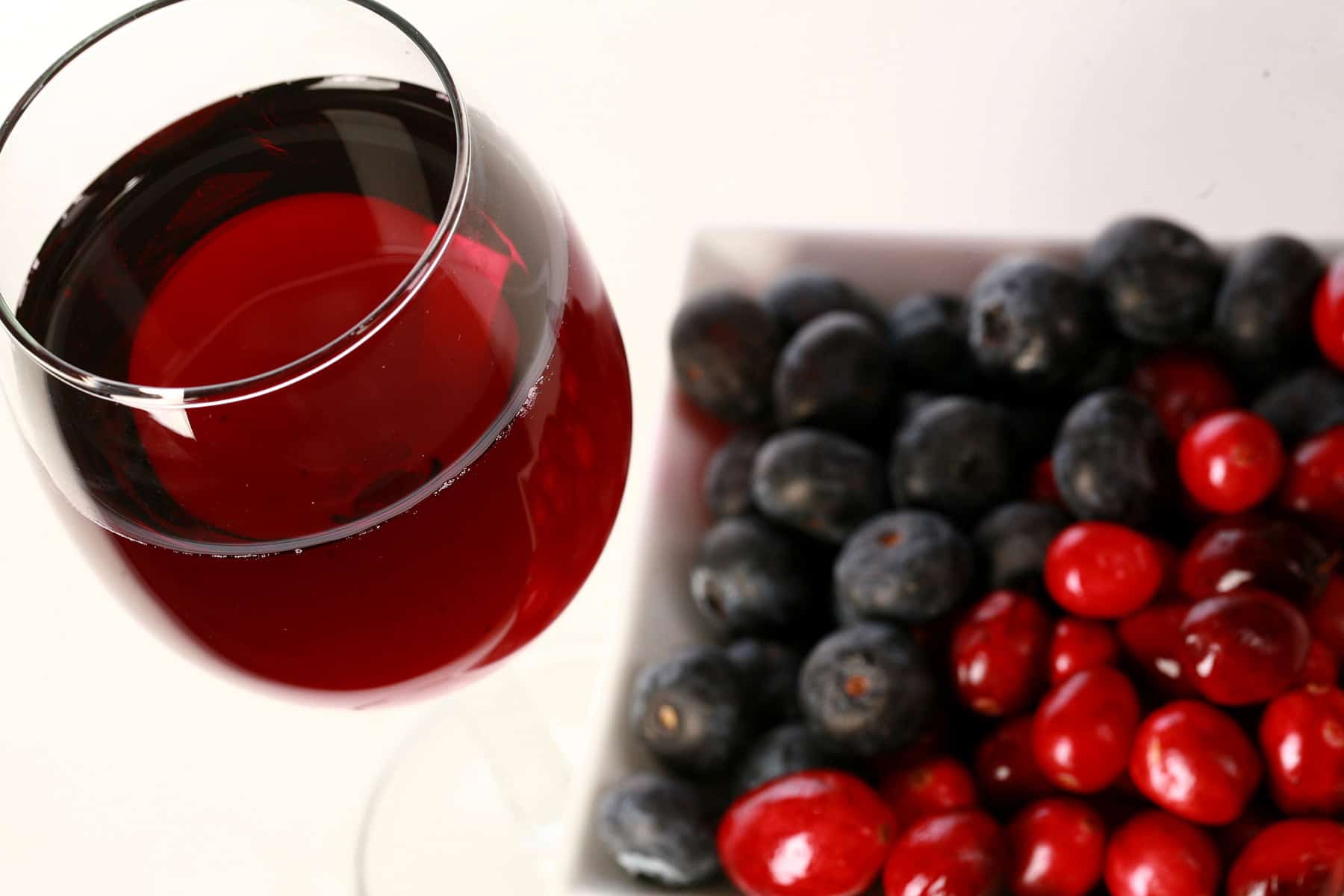 A glass of red wine is pictured next to a small bowl of cranberries and blueberries.