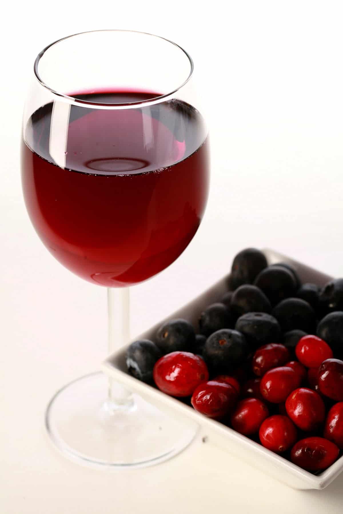 A glass of red wine - made from this faux lingonberry wine recipe - is pictured next to a small bowl of cranberries and blueberries