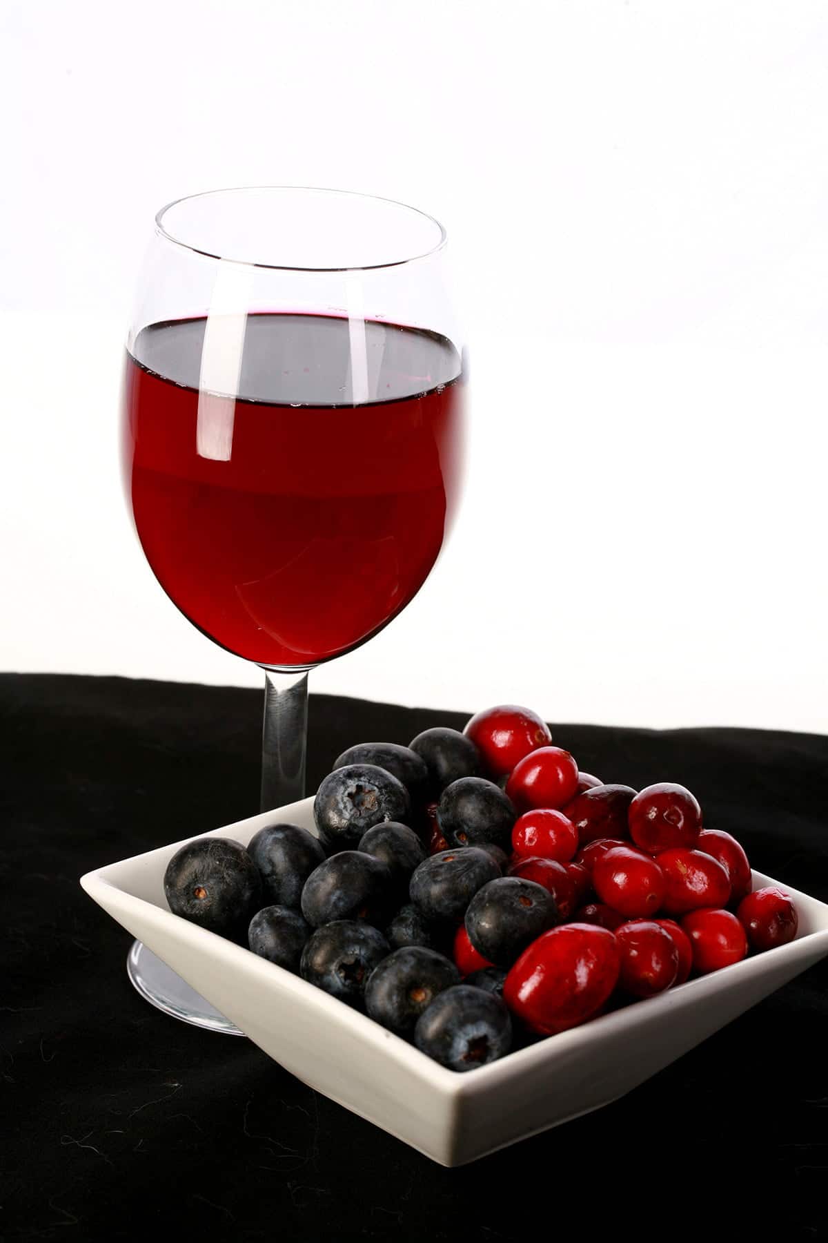 A glass of red wine - made from this faux lingonberry wine recipe - is pictured next to a small bowl of cranberries and blueberries