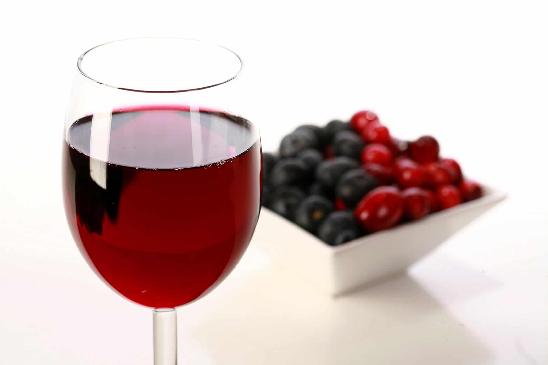 A glass of red wine is pictured next to a small bowl of cranberries and blueberries.