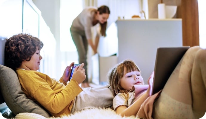 Enfants allongés qui utilisent des tablettes pendant que leur mère, en arrière-plan, range la cuisine.