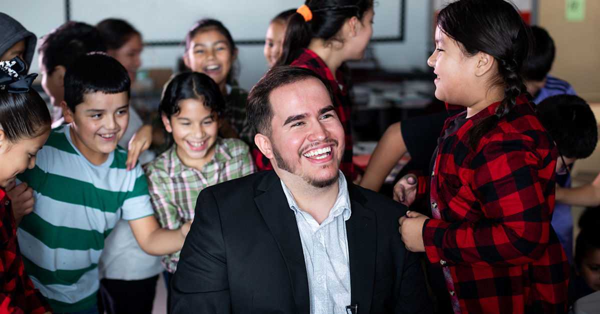 Uriel Iglesias, a 4th-grade bilingual teacher, sits in a chair as his student run up behind him for a group hug. All of them are smiling.