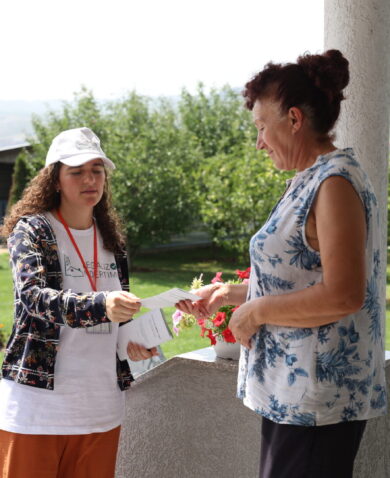 A young girl dressed in a white cap, white shirt, grey cardigan, and red pants hands an older woman, wearing a bun and blue and white floral top, a flyer.