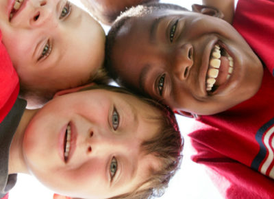 A group of elementary-aged boys huddled shoulder-to-shoulder, looking down at a camera that sits between them. They have huge smiles on their faces.