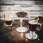 Gorgeous coffee glasses filled with hot coffee and cocktail glass filled with coffee beans standing on dark ground, surrounded by coffee beans.