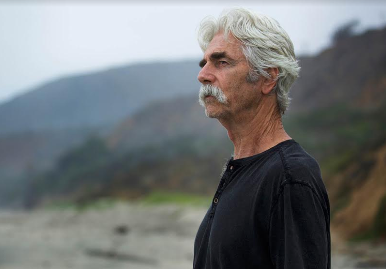 A man with a mustache, going gray, stands on a beach.