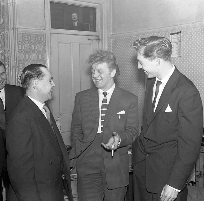 Three men in suits converse and laugh, taking life less seriously in a room with patterned wallpaper and wooden detailing. A portrait of another man is artfully visible above a door in the background.