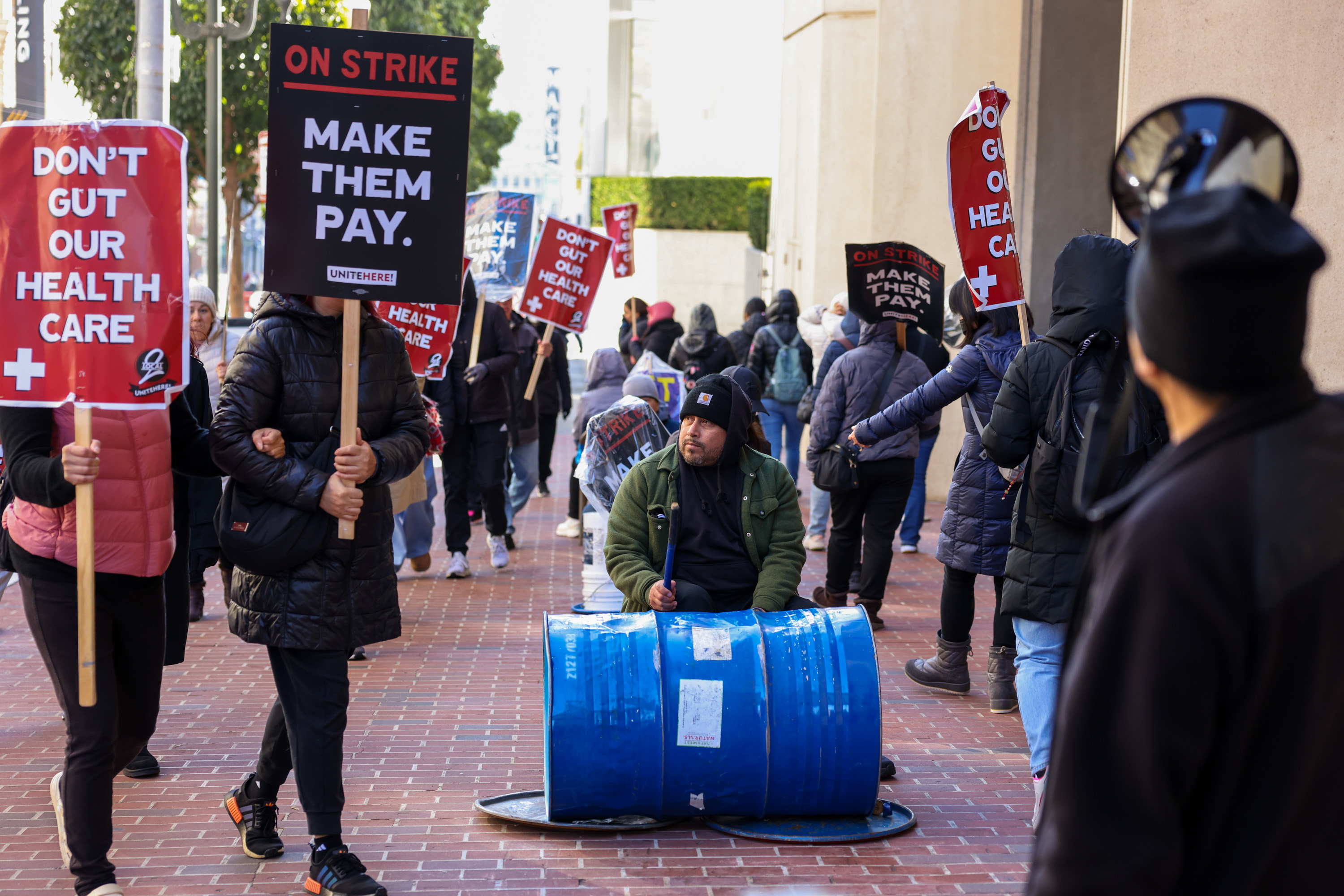 Protesters stand on a brick sidewalk holding signs that say "DON'T GUT OUR HEALTH CARE" and "MAKE THEM PAY," with a man drumming on a blue barrel.