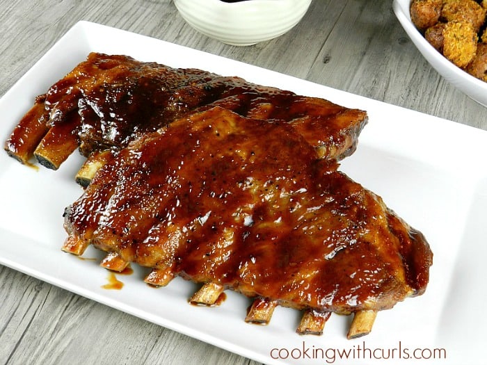 barbecue ribs on a white platter with barbecue sauce and a bowl of potatoes in the background