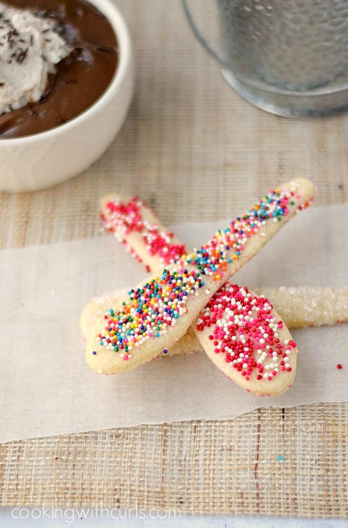 sprinkle covered pie crust spoons laying on parchment paper with a bowl of chocolate pudding in the upper left corner