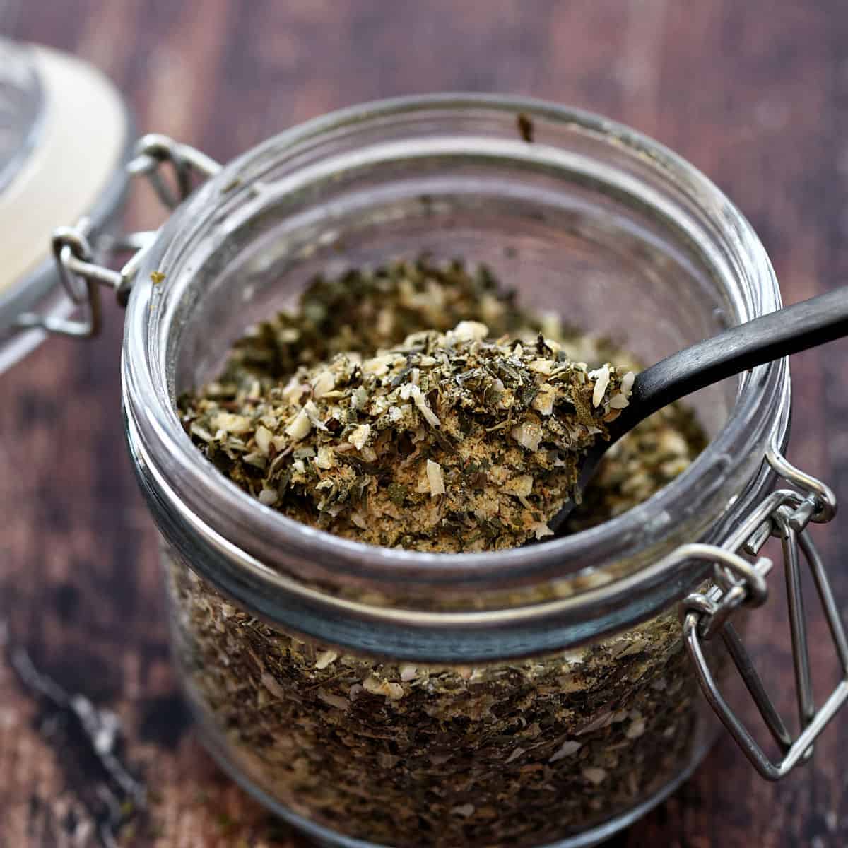 A small glass jar filled with Greek herbs and spices.