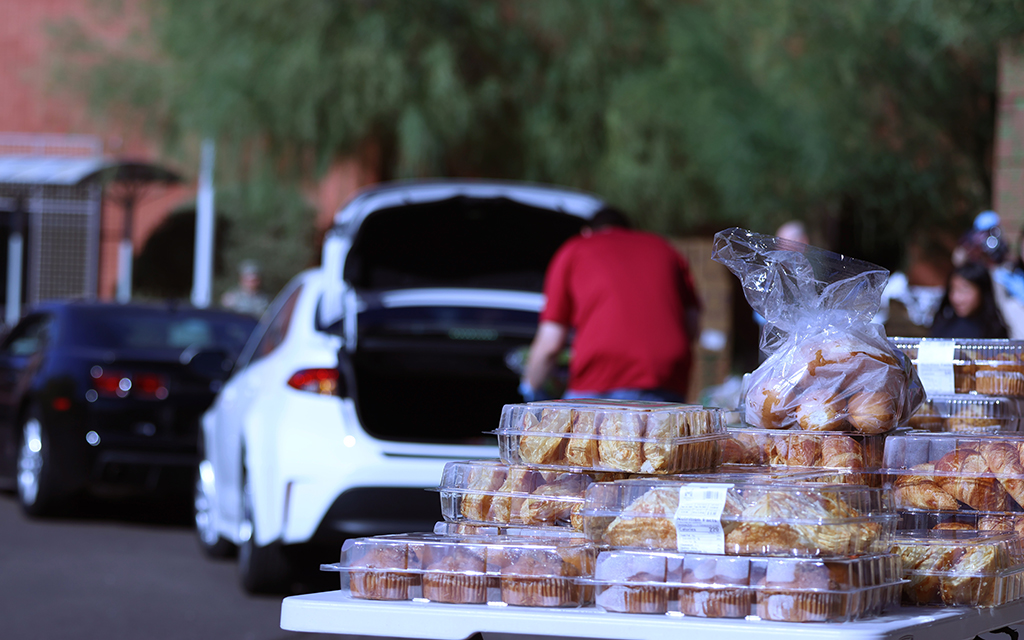 Table with containers of pastries in front of an open car trunk with a person nearby.