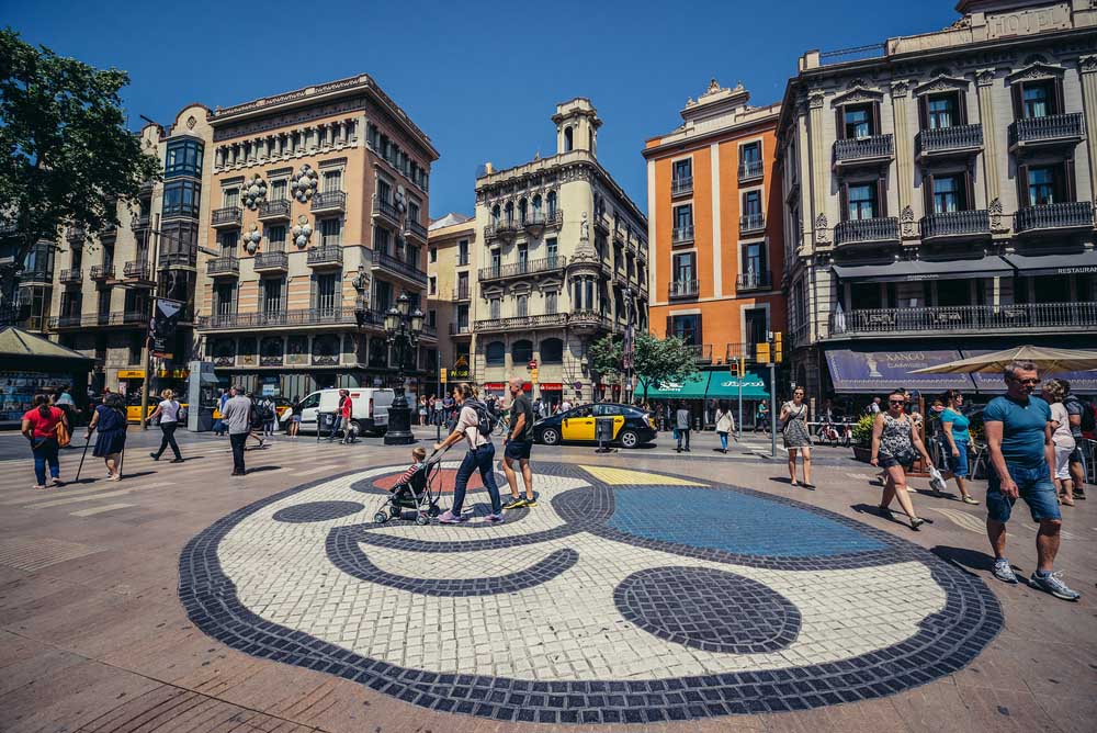 People walks at famous La Rambla Street, Pla de la Boqueria. View with mosaic created by Joan Miro and House of Umbrellas. Photo: Shutterstock