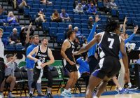 Group of Daemen Basketball team members celebrating on the basketball court with some fans sitting in seats behind them.