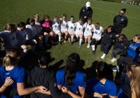 A circle of women soccer players kneeling on grass, arm-in-arm, with coaching staff standing behind them.