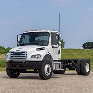 The Freightliner M2 106 Plus truck on a road in front of a green field.