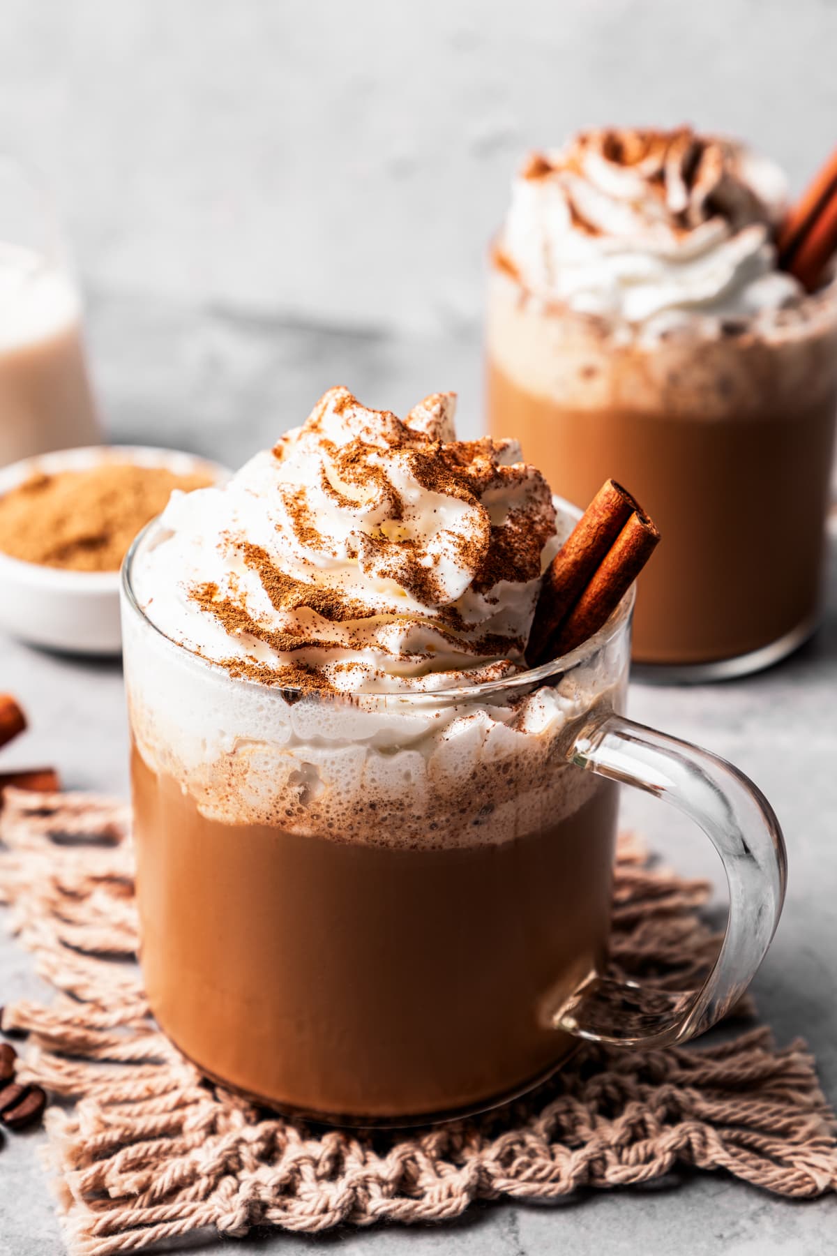 A cinnamon dolce latte in a glass mug garnished with whipped cream and a cinnamon stick, dusted with cinnamon, with a second latte in the background.