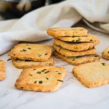 A stack of square cottage cheese crackers with green herbs rests on a white surface next to a white cloth. Some crackers are spread around in the foreground.