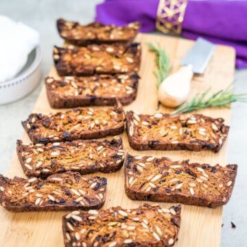 Slices of nut-and-seed bread arranged on a wooden board with a sprig of rosemary and a small cheese knife nearby.