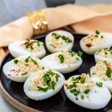 A plate of deviled eggs garnished with chopped chives and paprika, with a beige napkin in the background.